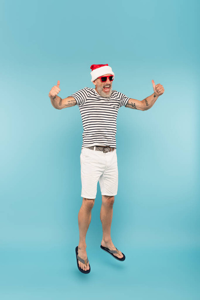 full length of happy man in sunglasses and santa hat jumping while showing thumbs up on blue - Photo, Image