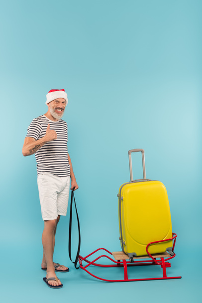 longitud completa de hombre feliz en sombrero de santa que muestra el pulgar hacia arriba mientras está de pie cerca de equipaje en trineo en azul - Foto, Imagen