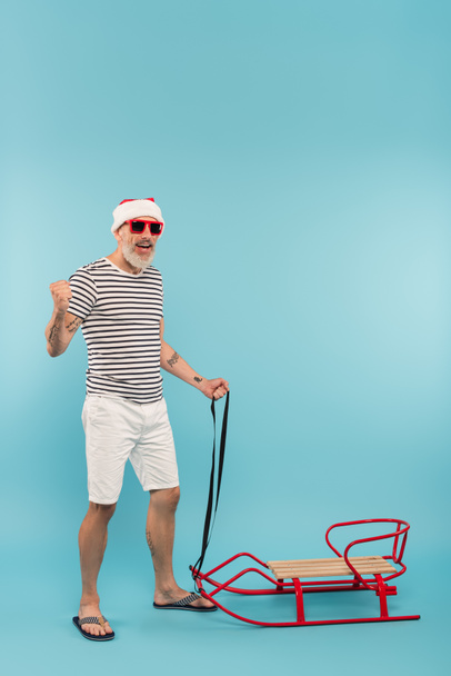 повна довжина щасливого чоловіка в капелюсі Санта і сонцезахисних окулярах, що стоять поруч з санчатами на синьому
 - Фото, зображення