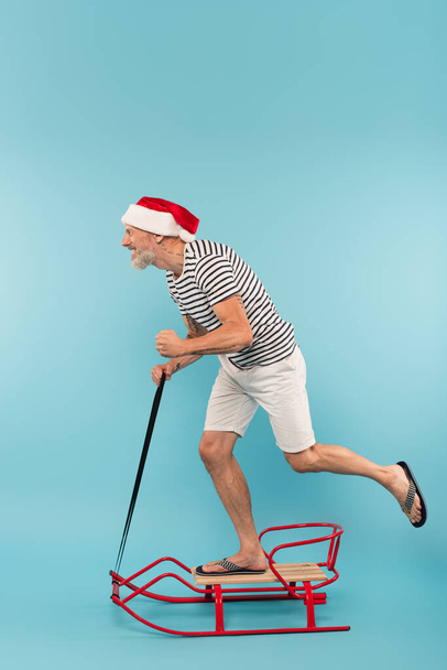 Seitenansicht eines fröhlichen Mannes mit Weihnachtsmannhut, der auf einem Schlitten auf blau reitet - Foto, Bild