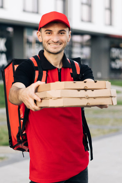 Attractive young delivery man courier in red cap smiling into camera holding pizza boxes delivering fast food around city. Outdoor portrait. Urban worker. Concept of courier, home delivery, pizza - Photo, Image