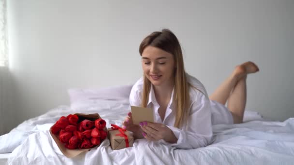 Boldog szülinapot! Boldog szőke lány fekszik reggel az ágyon egy csokor virággal és egy ajándék elővesz egy képeslapot a felirattal. - Felvétel, videó