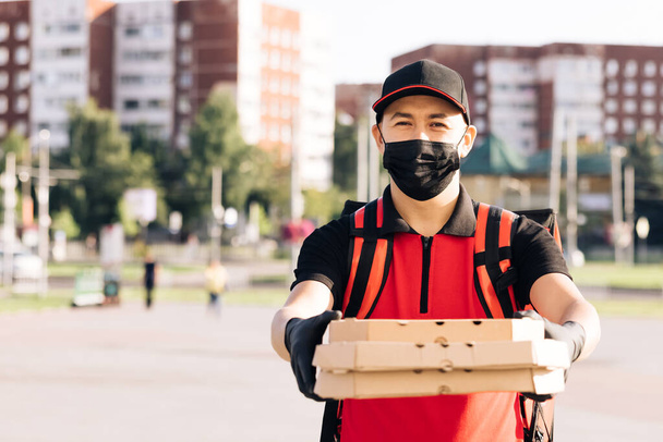 Young male food courier wearing protective mask while walking down city street with insulated red backpack delivering order. Delivery services. Excellent delivery - 写真・画像