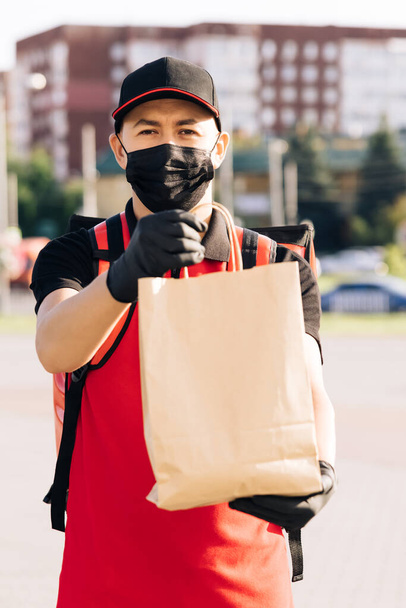 Food delivery man holding paper bag with food at street outdoors home, deliveryman in protective medical face mask gloves, coronavirus COVID-19 epidemic quarantine outbreak. Delivery services - Photo, Image