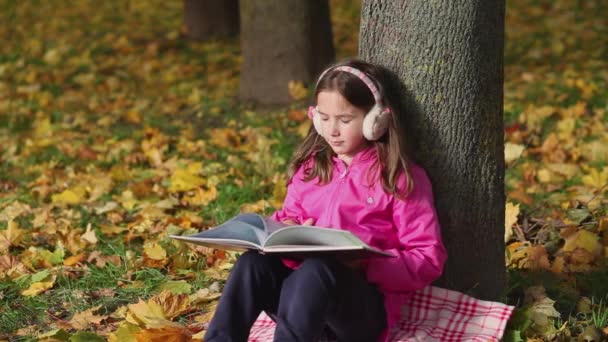 Χαριτωμένο κοριτσάκι διαβάζει ένα βιβλίο που κάθεται κάτω από ένα δέντρο. Πολύχρωμα φύλλα πέφτουν από το δέντρο. - Πλάνα, βίντεο