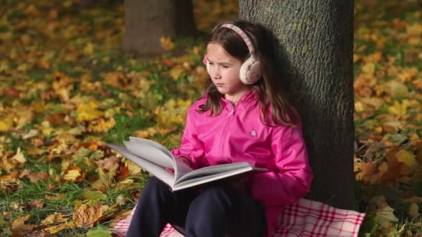 Χαριτωμένο κοριτσάκι διαβάζει ένα βιβλίο που κάθεται κάτω από ένα δέντρο. Πολύχρωμα φύλλα πέφτουν από το δέντρο. - Πλάνα, βίντεο