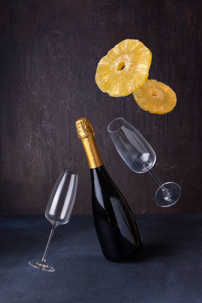 бутылка игристого вина, бокалы, ананасы летают на темном фоне - Фото, изображение