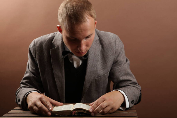 Przystojny, pewny siebie biznesmen w garniturze, siedzący samotnie na brązowym tle i czytający książkę. Obok drewnianego stołu - Zdjęcie, obraz