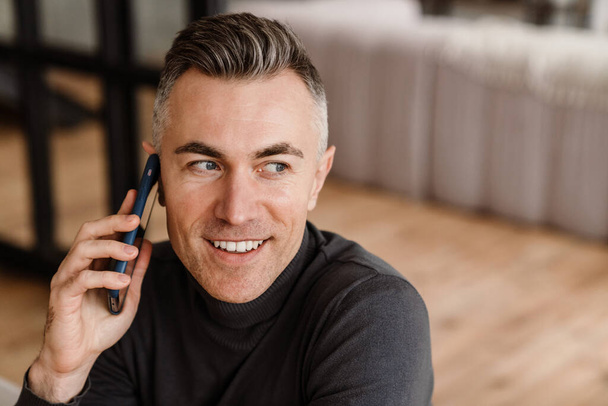 Красивый счастливый мужчина улыбается и разговаривает по мобильному телефону дома - Фото, изображение