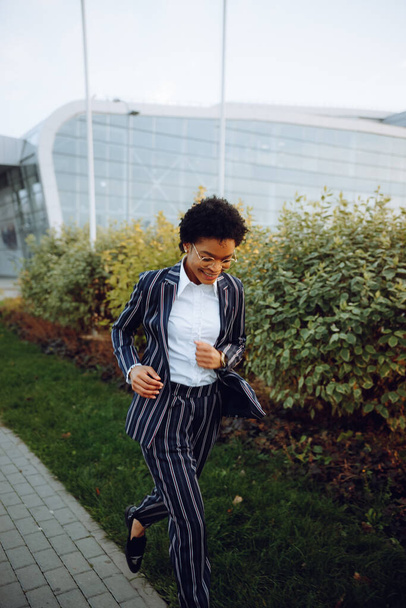 4k. Travel, Digital. A charming African American woman in an elegant striped suit, looks at the camera, running, smiles and feels fantastic. She run against the bushes near the airport. Businesswoman - Photo, image