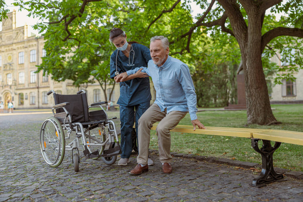 Full length shot of supportive young nurse wearing face shield and mask helping mature man, recovering patient in wheelchair to sit on the bench in the park near hospital - Fotó, kép