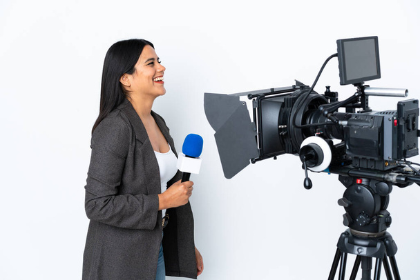 Δημοσιογράφος Κολομβιανή γυναίκα που κρατάει μικρόφωνο και μεταδίδει ειδήσεις σε λευκό φόντο γελώντας στην πλαϊνή θέση - Φωτογραφία, εικόνα