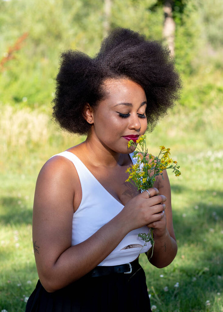An African American female smelling the yellow wildflowers while on picnic at Tormes River in Salamanca City, Spain - Foto, imagen