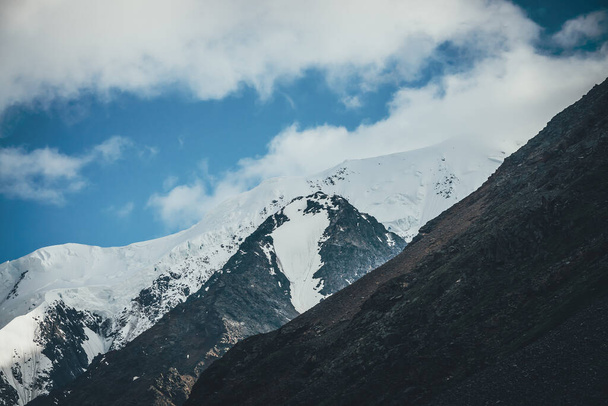 Огромный горный ландшафт с большой снежной вершиной. Атмосферный пейзаж с белоснежной высокой горой с ледником. Большие горы со снегом. Живописный вид на гигантские черные скалы с белым снегом. - Фото, изображение