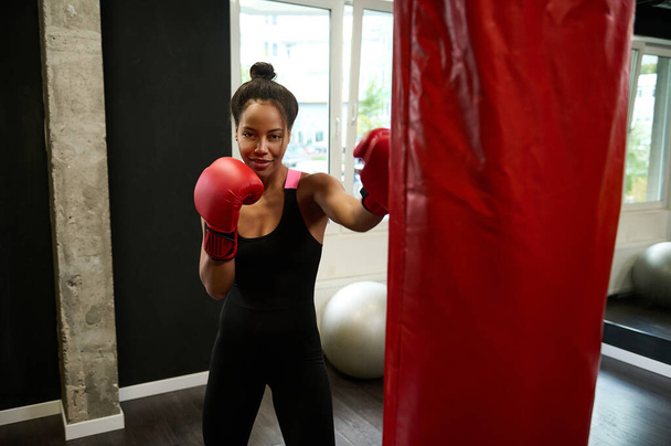 Belle boxeuse athlète africaine portant des gants de boxe rouges frappe sur le sac de boxe, regarde la caméra tout en effectuant des arts martiaux de combat à la salle de sport. Mode de vie sain et actif concept - Photo, image