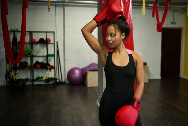 Portrait de belle femme sportive africaine, jolie boxeuse en vêtements de sport noirs et gants de boxe rouges, regardant sur le côté, sourit mignon appuyé sur un sac de boxe au gymnase de boxe. - Photo, image