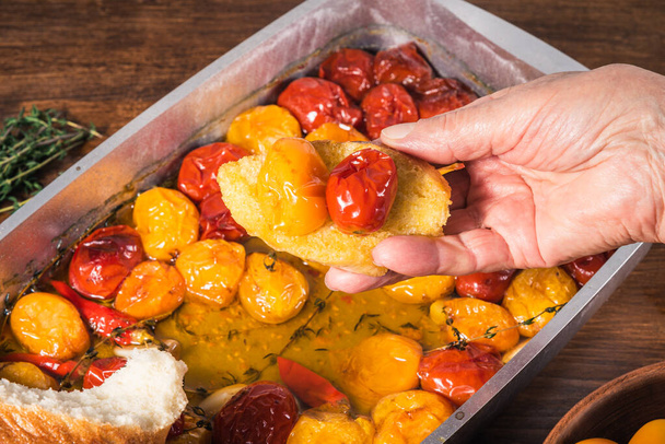 Frau probiert Confit aus roten und gelben Kirschtomaten, Hand einer älteren Frau mit einem Stück Baguette und gebackenen Tomaten aus nächster Nähe - Foto, Bild