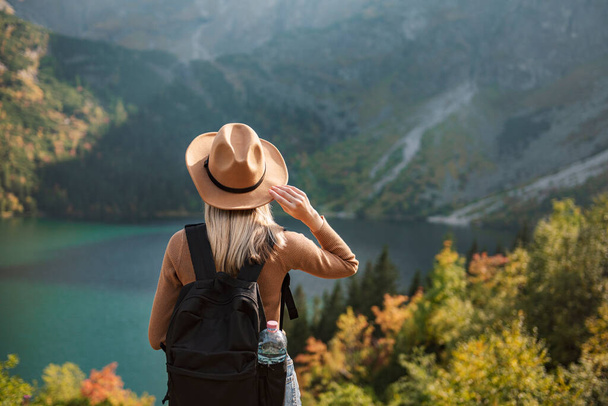 Woman traveler wearing hat and looking at amazing mountains and lake, wanderlust travel concept. Lake Morskoy Eye in the Polish Tatras. - Valokuva, kuva