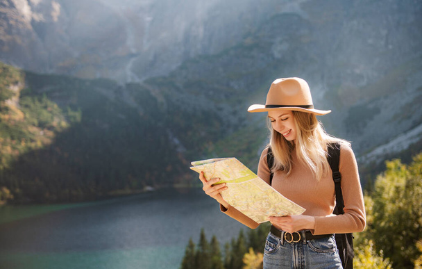 欲望と旅行の概念。帽子のスタイリッシュな旅行者の女の子は地図を見て、森を探索します。森の中の湖を旅するリュックを持つ若い女性.  - 写真・画像