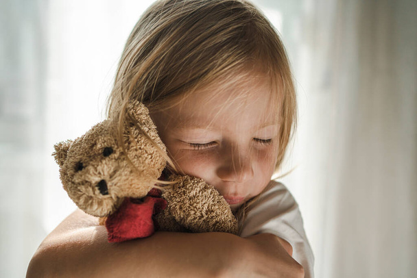 Little Girl with Blond Hair Gently Hugs her Favorite Soft Toy. Girl With a Teddy Bear. Teddy Bear and Kid - Φωτογραφία, εικόνα