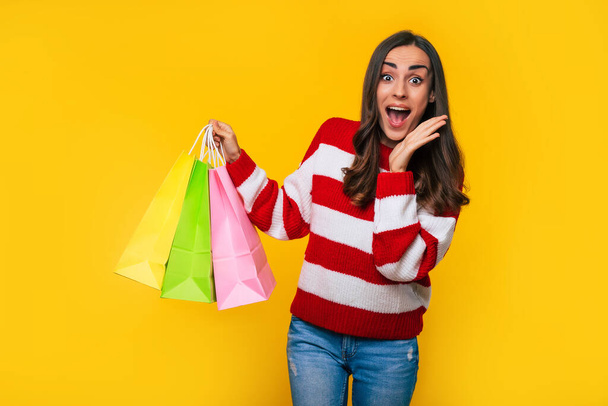 Close up photo of cute excited woman in a striped sweater with many colorful shopping bags in hands isolated on yellow background - Foto, afbeelding