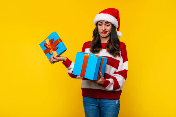 Young disappointed cute girl in Christmas hat is unpacking gift box on yellow background with unhappy face - Foto, Imagem