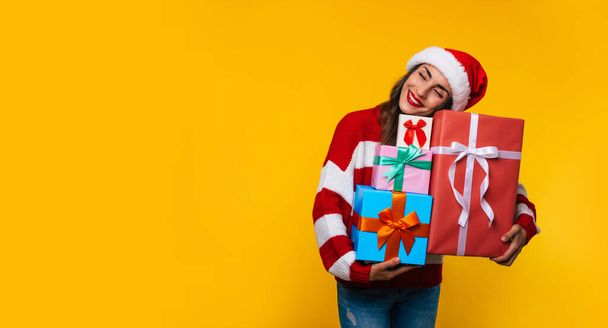 Gorgeous smiling woman with many cool Christmas gift boxes in hands while she is having fun and posing on yellow background  - Фото, изображение