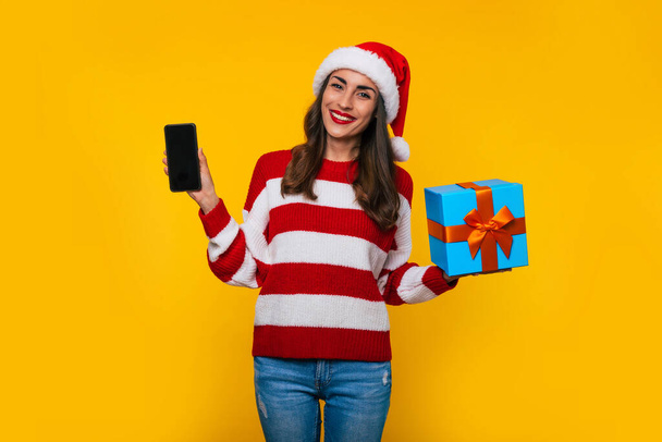 Young beautiful excited smiling woman while she shows Christmas gift box and smart phone in hands and posing on yellow background  - Foto, Imagem