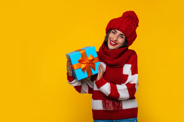 Nahaufnahme Foto von schönen aufgeregt lächelnde Frau in Winterkleidung mit Weihnachtsgeschenk-Box in den Händen hat Spaß beim Posieren auf gelbem Hintergrund  - Foto, Bild