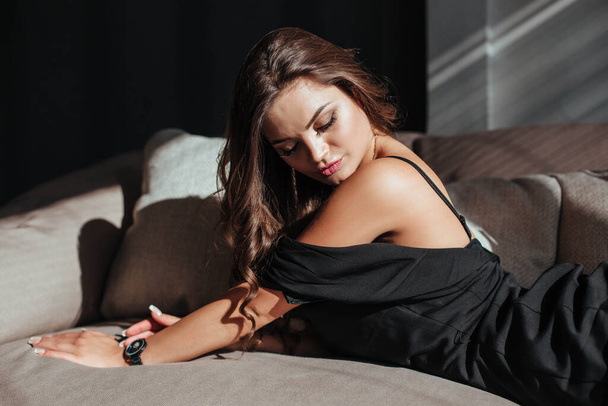Portré egy szexi barnáról fekete ruhában, sötét háttérrel. lány leveszi a ruháját lassan a hálószobában - Fotó, kép