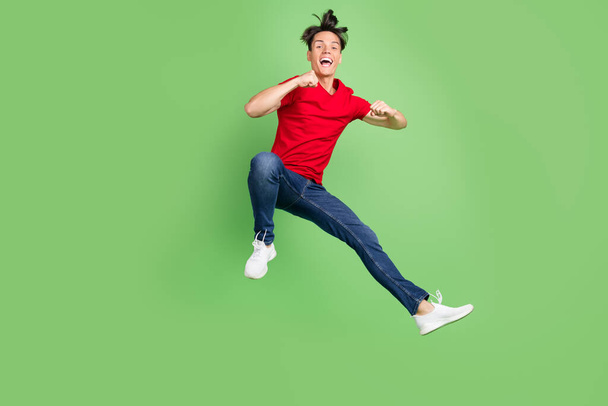 Photo of funny excited young guy dressed red t-shirt jumping high rising fists smiling isolated green color background - Фото, изображение
