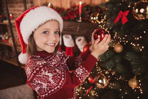 Photo of dreamy pretty schoolgirl dressed red sweater headwear decorating christmas tree smiling indoors room home house - Φωτογραφία, εικόνα