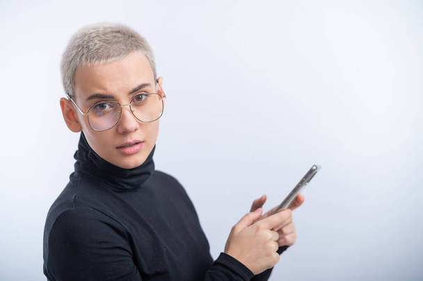 Young caucasian woman with short hair uses a smartphone on a white background. - Foto, afbeelding