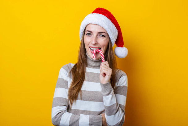 Sourire jeune femme dans le chapeau du Père Noël manger des bonbons de Noël sur fond jaune. - Photo, image