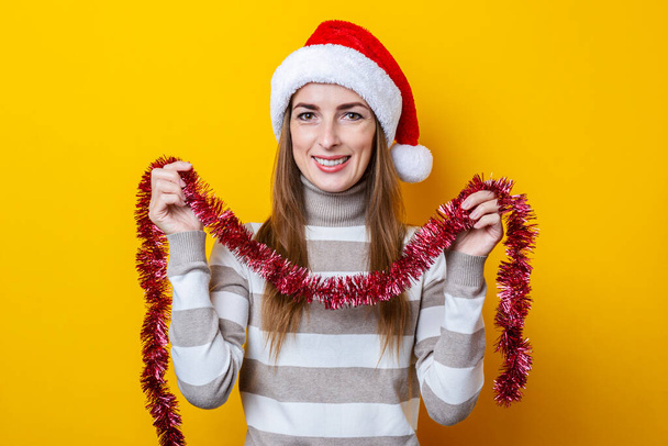 Lächelnde junge Frau mit rotem Weihnachtslametta zur Dekoration auf gelbem Hintergrund.  - Foto, Bild