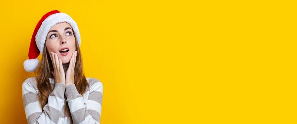 Pensive surprised young woman in Santa Claus hat on a yellow background. Banner. - Foto, afbeelding