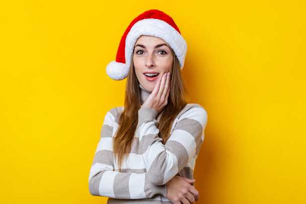 Überraschte junge Frau mit Nikolausmütze auf gelbem Hintergrund. - Foto, Bild