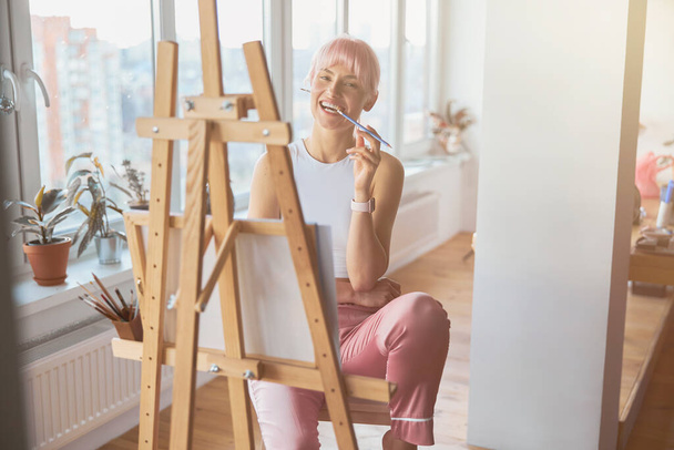 Θετική γυναίκα με πολύχρωμα τσιμπήματα μαλλιών μολύβι κάθεται στο καβαλέτο - Φωτογραφία, εικόνα