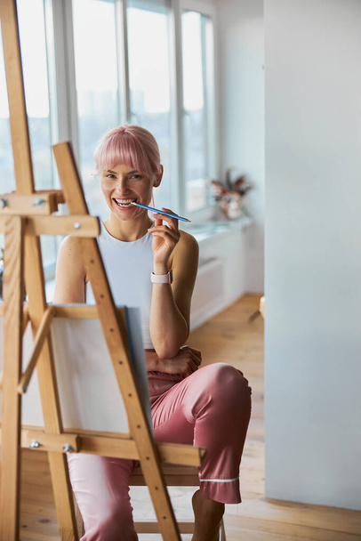 Attractive woman with pink hair bites pencil at easel on balcony - Foto, Imagem