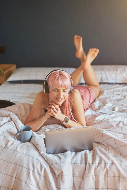 Красивая женская модель с наушниками и чашкой смотрит на экран современного ноутбука на кровати - Фото, изображение