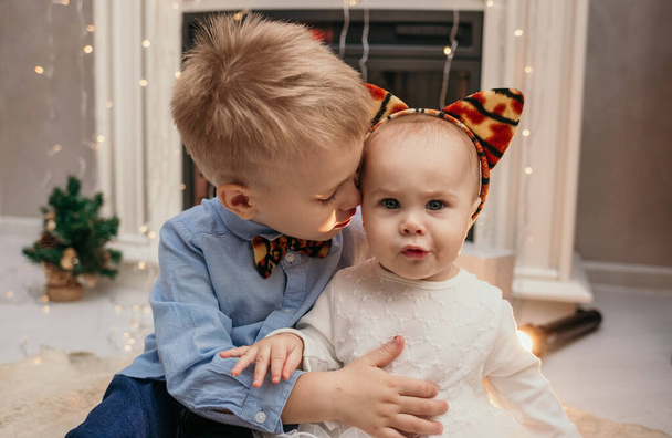 irmão e irmã em roupas festivas estão sentados no tapete perto da lareira na sala - Foto, Imagem