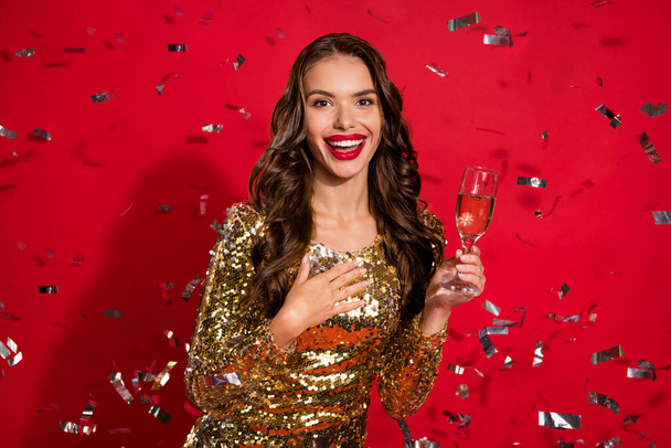 Foto von dankbar schöne Dame trinken Champagner Hand Brust fallen Konfetti tragen goldenes Kleid isoliert rote Farbe Hintergrund - Foto, Bild