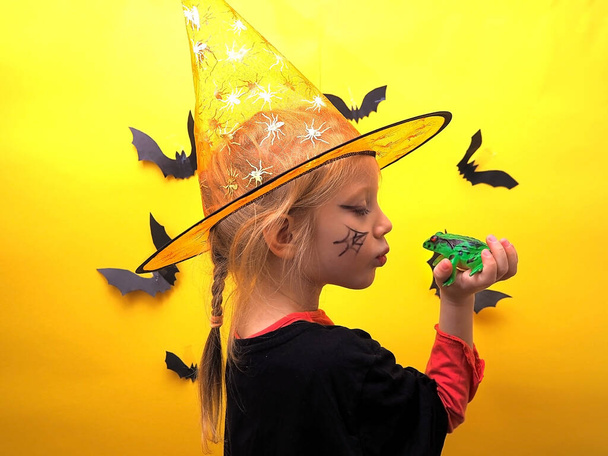 Ein Mädchen im Hexenkostüm auf einem Hintergrund mit Fledermäusen küsst einen Frosch. Kindermädchen als Hexe auf dem Hintergrund von Fledermäusen verkleidet. Vorbereitungen für Halloween. Halloween-Kostüm. - Foto, Bild