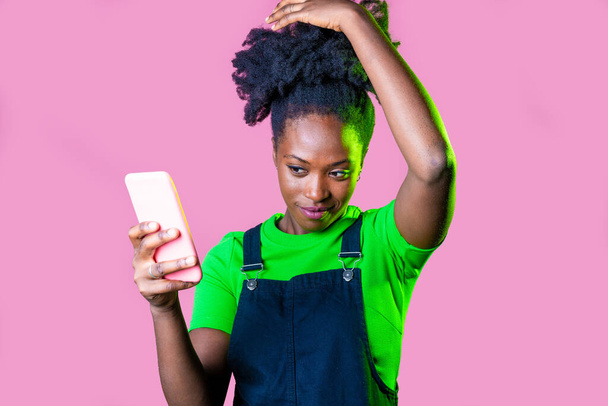 Jovem mulher negra usando smartphone videocalling tocando cabelo isolado fundo rosa - Foto, Imagem