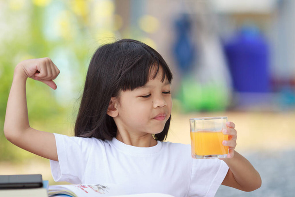 アジア系タイ人の子供の女の子明るい笑顔でかわいい顔,白いシャツを着て,健康.屋外に座ってテーブルの上に本やオレンジジュースのガラスがあります背景がぼやけている緑. - 写真・画像