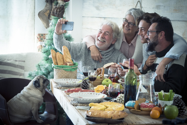 Hombre mayor tomando selfie utilizando el teléfono móvil con la familia en la mesa de comedor con perro de mascota sentado en la silla. Tres generaciones de familia celebrando la Navidad mientras cenan juntos. - Foto, Imagen