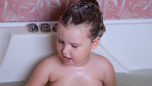 Mała zabawna dziewczynka 3-letnia kąpie się i myje z pianką na głowie w wodzie w łazience w domu. Koncepcja pielęgnacji ciała dziecka, higiena. - Zdjęcie, obraz