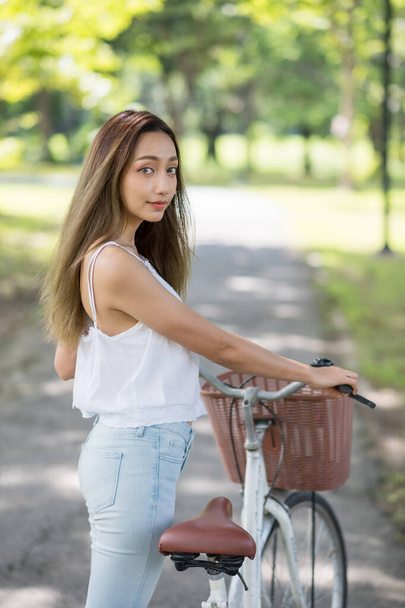 Retrato verbal de una joven asiática sonriente sosteniendo una bicicleta en el parque con follaje bokeh fondo en el parque de primavera. Personas Concepto de estilo de vida saludable y de ocio. - Foto, imagen