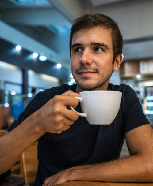 young hispanic man looks around with a smiling expression while having a coffee in a coffee shop - Foto, imagen