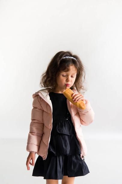 A cheerful brunette girl, dressed in a black dress and a pink squat, holds a baguette in her hands and eats. On white background. - Fotoğraf, Görsel
